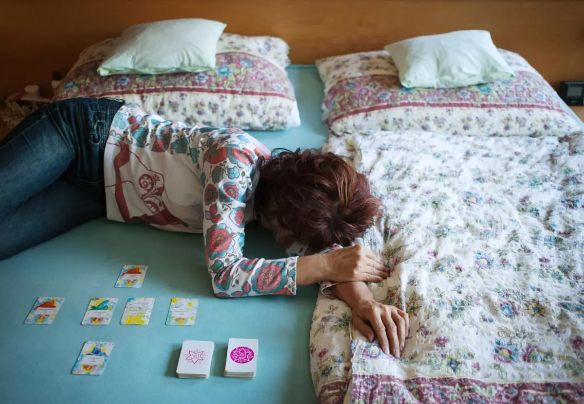 Person lying facedown on a teal bed sheet between two floral quilted beds, with colorful cards spread on the surface.