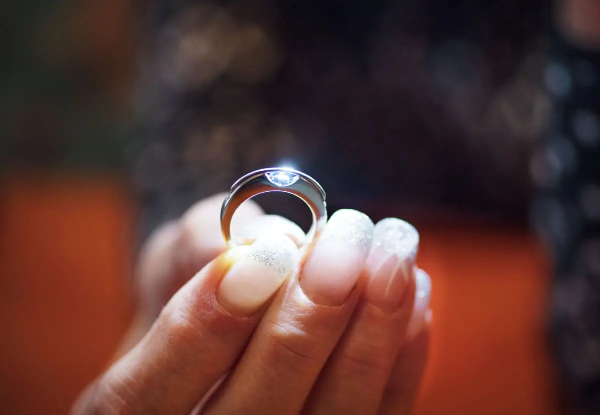 Close-up of a silver diamond ring held between fingertips against a dark, blurred background.