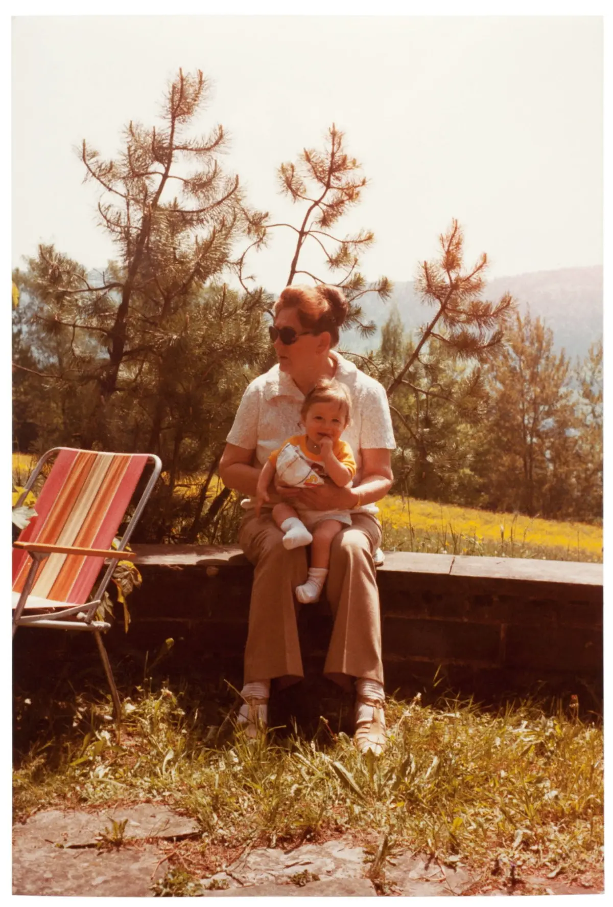 Elderly woman sitting on a low wall outdoors, holding a baby on her lap, with a striped chair and pine trees in the sunny yard.