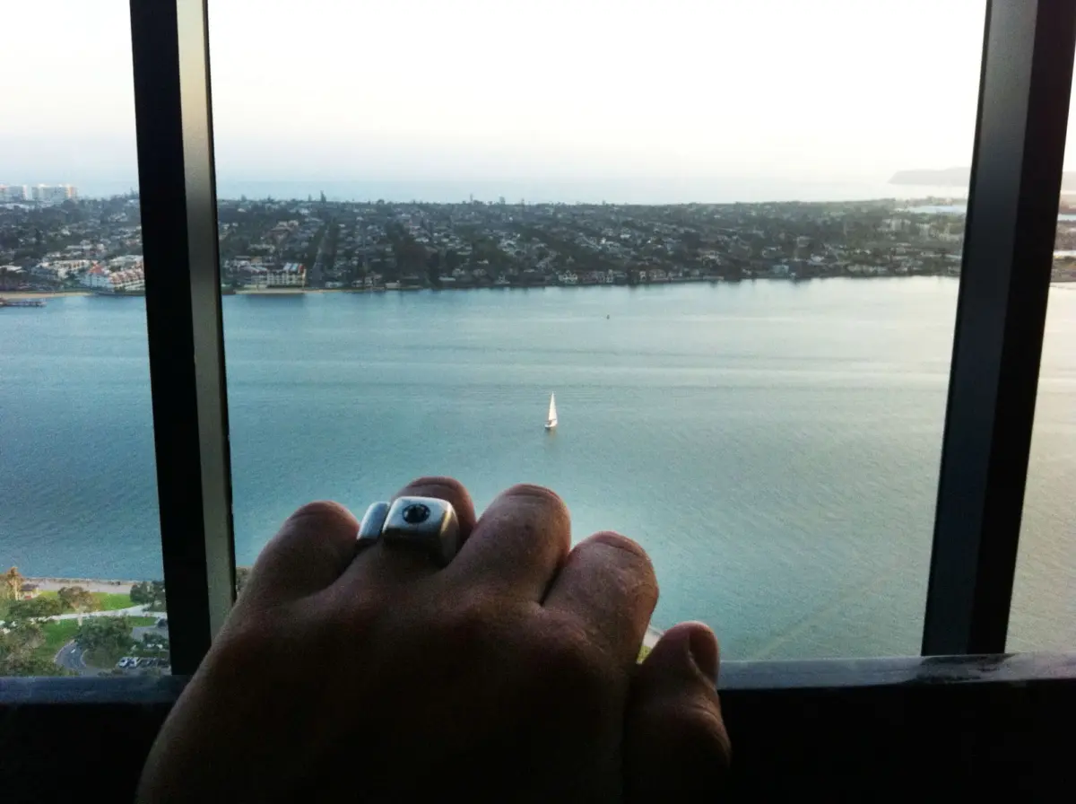Hand with ring holding a small camera seen through window frames, overlooking a coastal city and a sailboat on calm water.