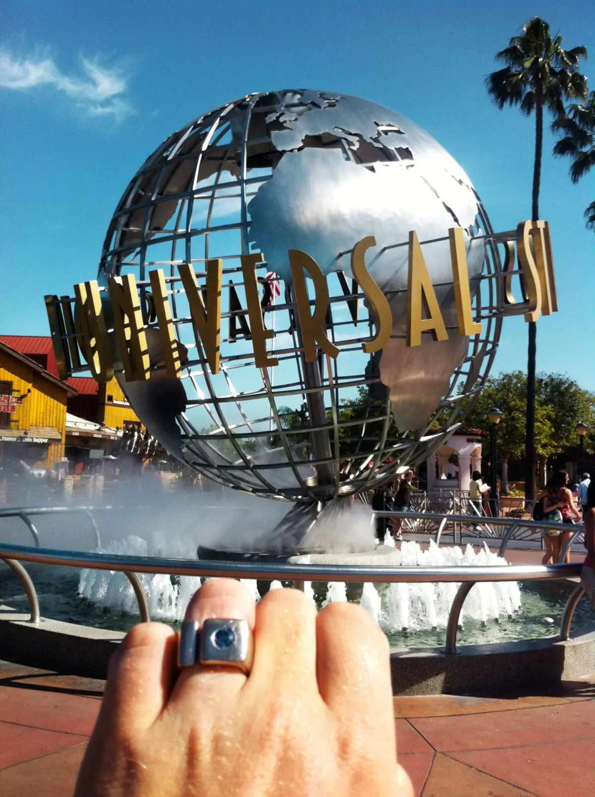 Metal globe sculpture with 'UNIVERSAL' letters, fountain around its base, palm trees, and a person taking a photo in a sunny plaza.
