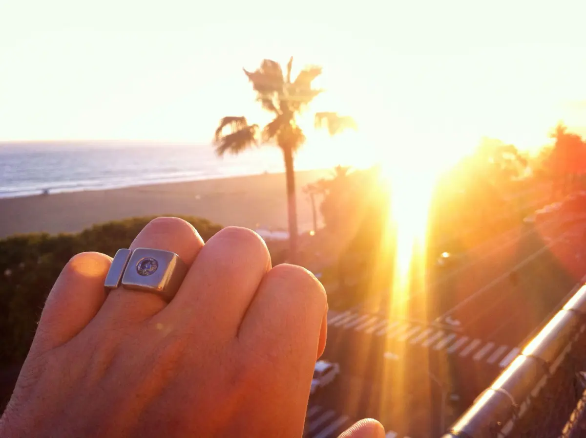 Close-up of a hand wearing a square-faced silver ring, held up against a sunlit beach with palm trees at sunset.