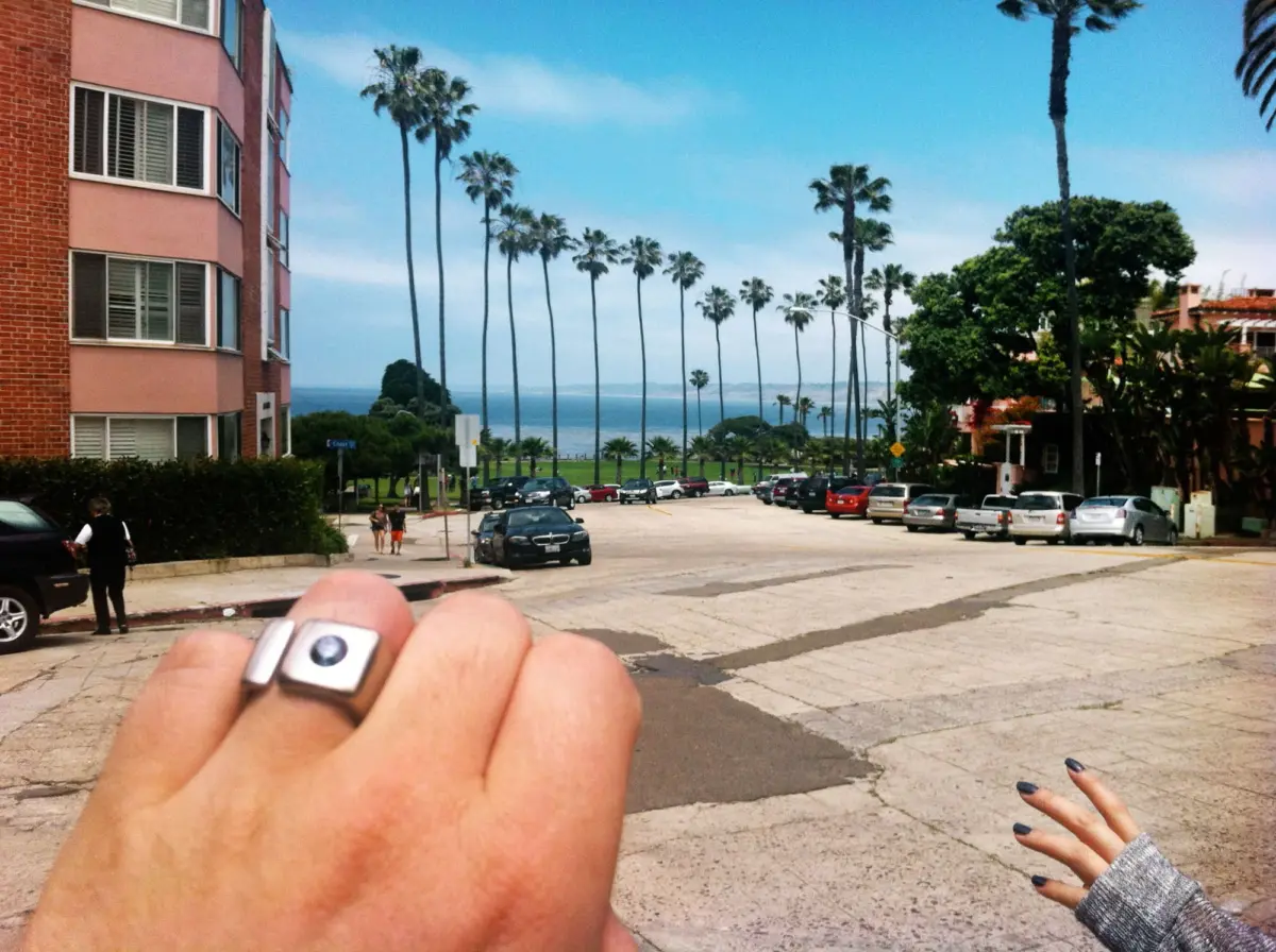 Close-up of a hand with a chunky silver ring and small device; palm-lined street, parked cars, and the ocean visible in the background.