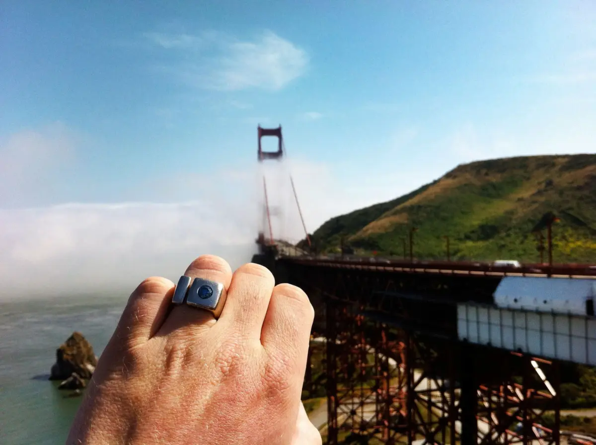 Close-up of a hand wearing a silver ring with a blue gem, with the Golden Gate Bridge and hills in the background.