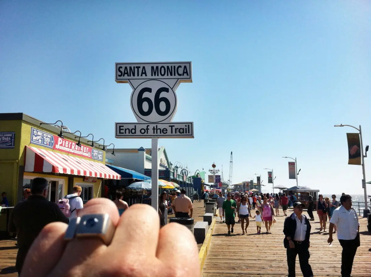 Santa Monica Route 66 End of the Trail sign on a busy boardwalk with colorful shops and pedestrians.