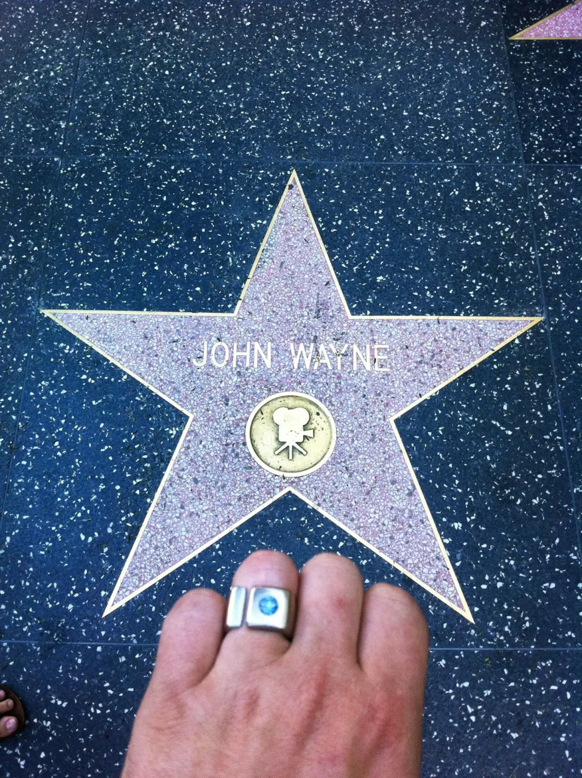John Wayne star on Hollywood Walk of Fame; pink terrazzo star on speckled black pavement, foreground shows a hand wearing a silver ring.