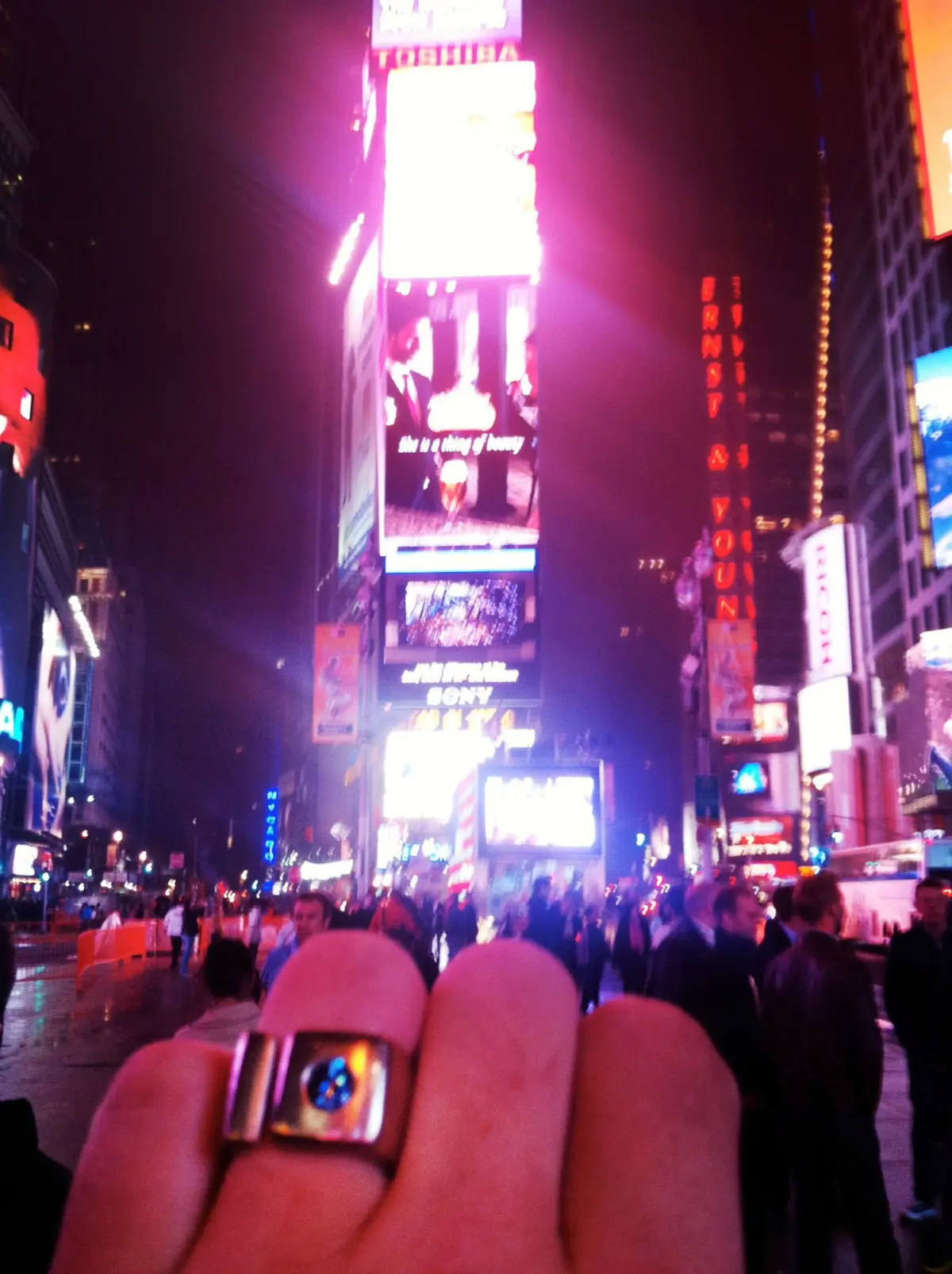 Close-up of a hand holding a smartphone in the foreground, crowded street with neon billboards in Times Square at night.