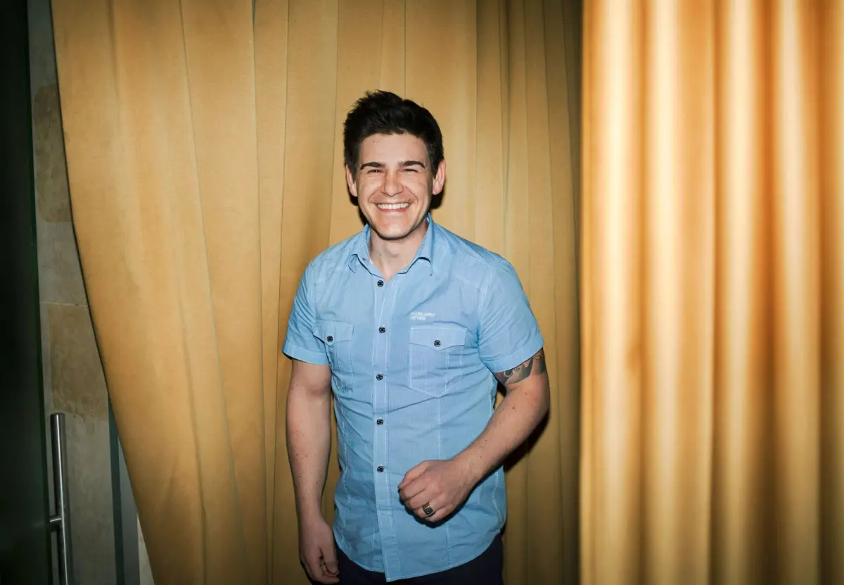 Smiling person in a light blue short-sleeve shirt stands in front of beige curtains.