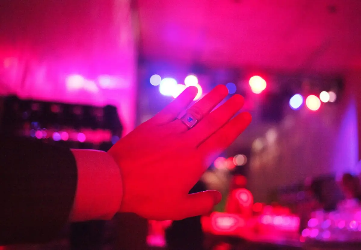 Raised hand with silver ring in foreground; pink and purple neon bokeh from a nightclub in the background.