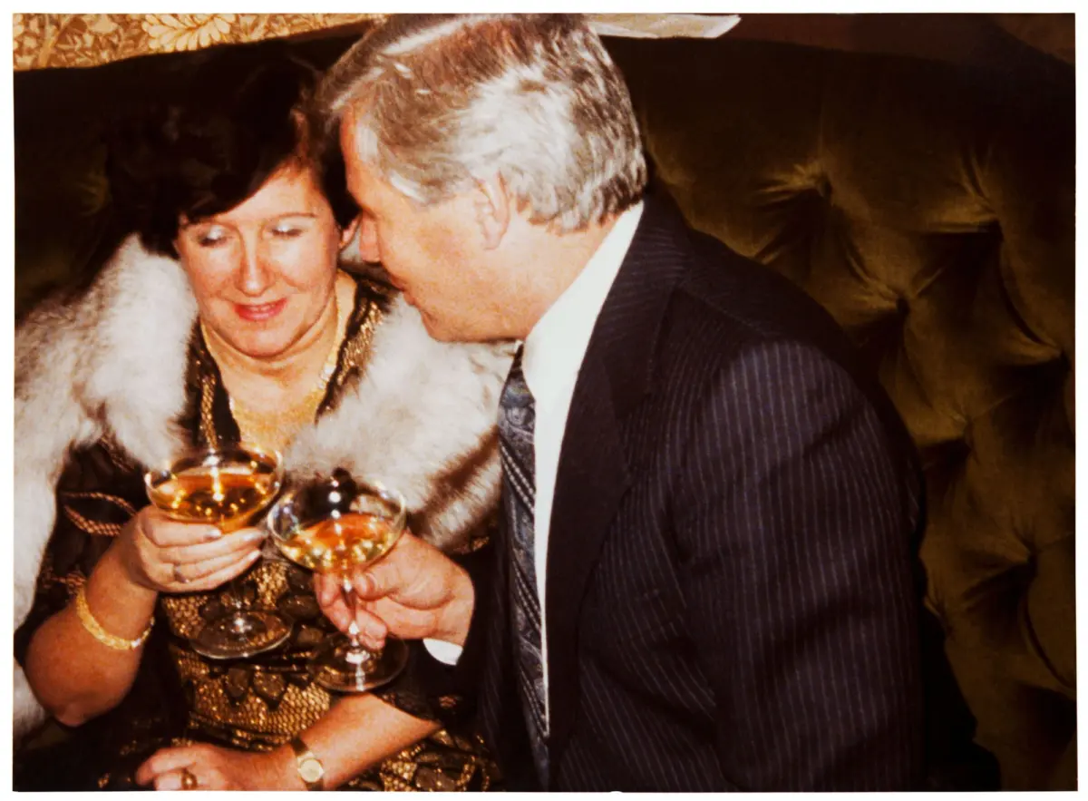 Elderly couple toasting with champagne glasses at a formal indoor party, woman in fur stole and gold jewelry, man in pinstripe suit.