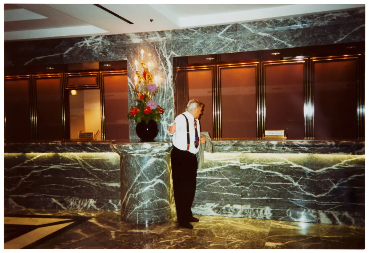 Older man in white shirt and suspenders stands holding a coat in a marble hotel lobby beside elevator doors and a floral arrangement.