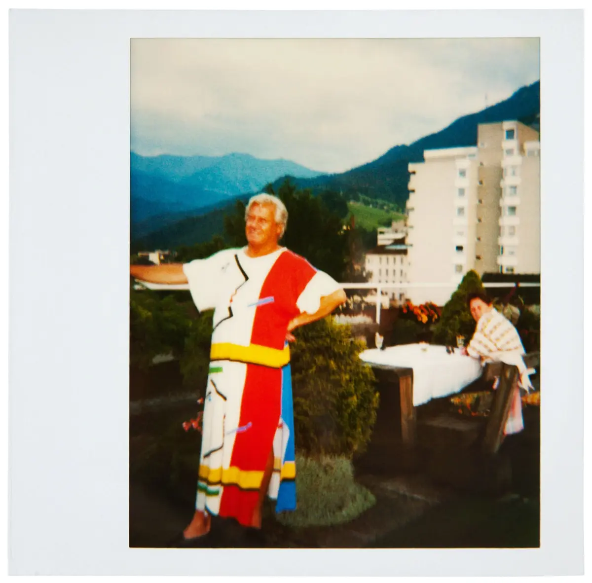 Adult person in a color-blocked outfit stands with arms outstretched outdoors, with apartment buildings and hills in the background.