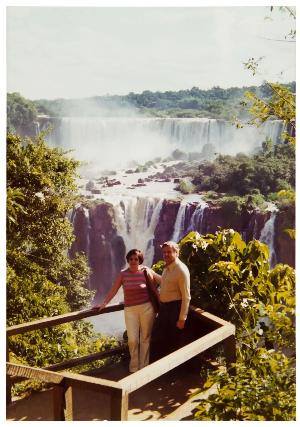 Two tourists pose on a wooden railing overlooking a large tropical waterfall with mist and lush jungle.
