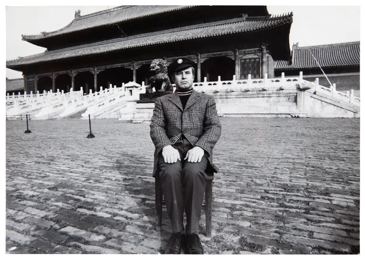 Man in plaid blazer and cap sits on a chair in front of a Chinese imperial palace, with a guardian lion statue and stone steps.