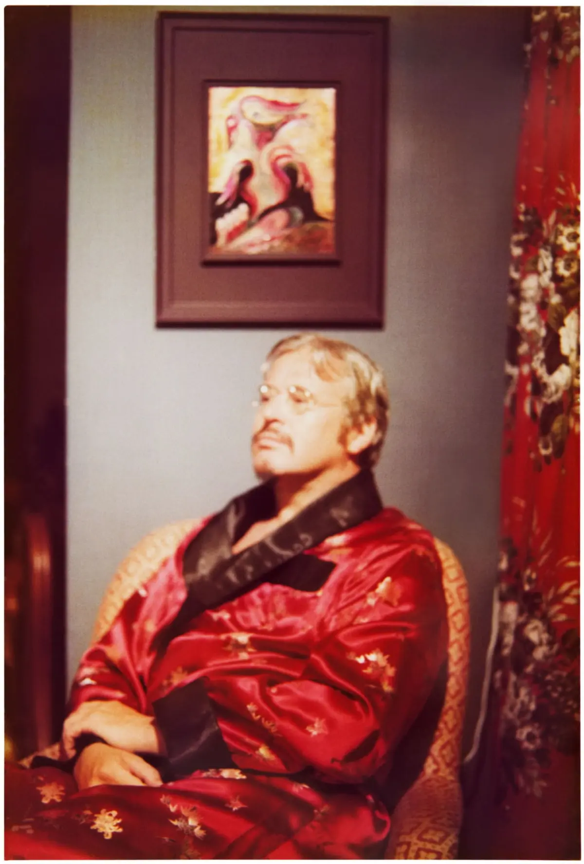 Elderly man wearing a red satin robe sits in an ornate chair, looking upward; framed artwork on gray wall behind, floral curtains to the side.