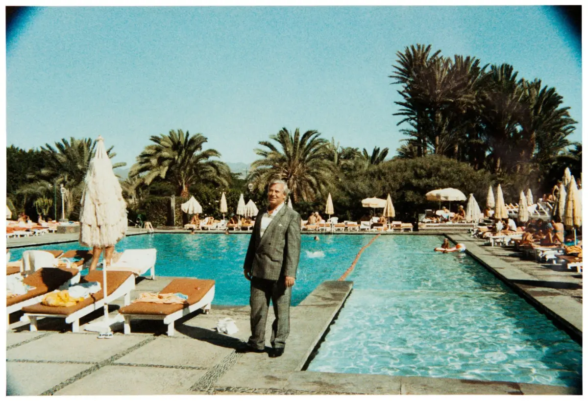 Person in gray suit stands poolside at a palm-lined resort, with lounge chairs and umbrellas around the pool.