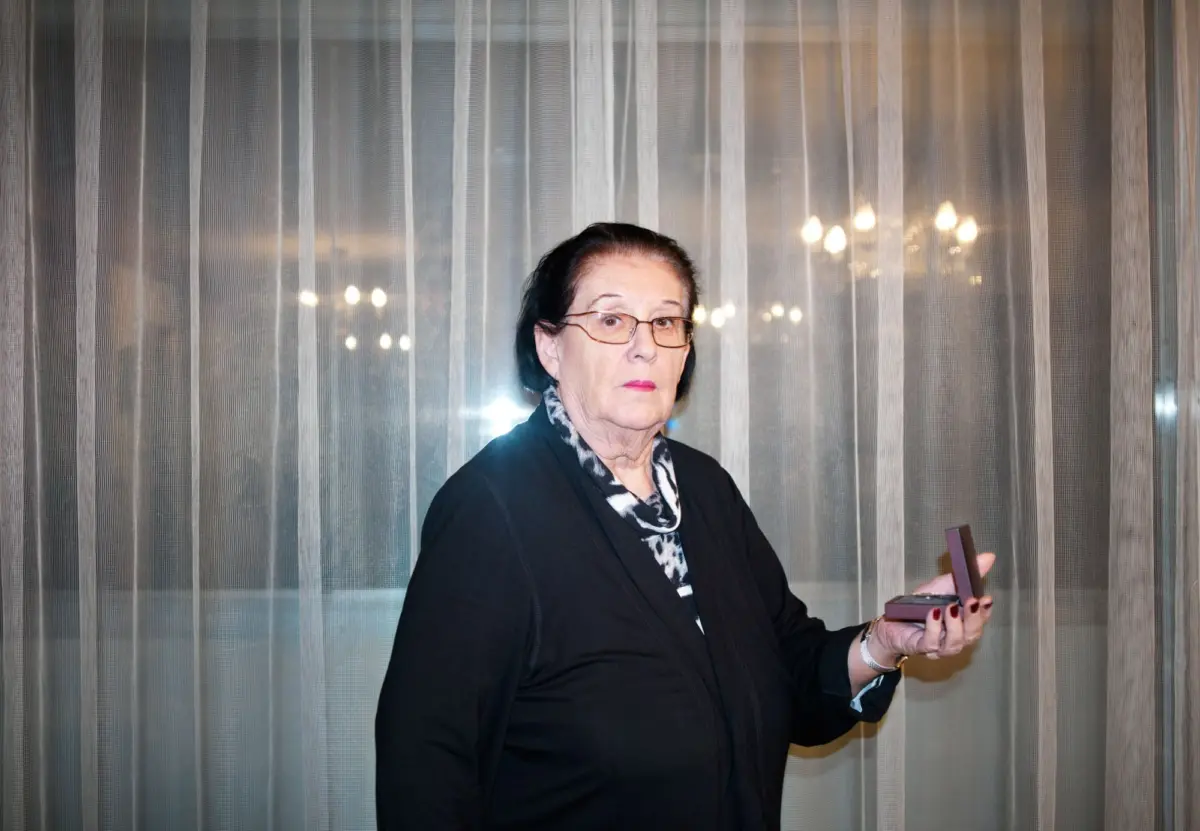 Older woman with glasses holding an open jewelry box, standing in front of sheer vertical blinds.
