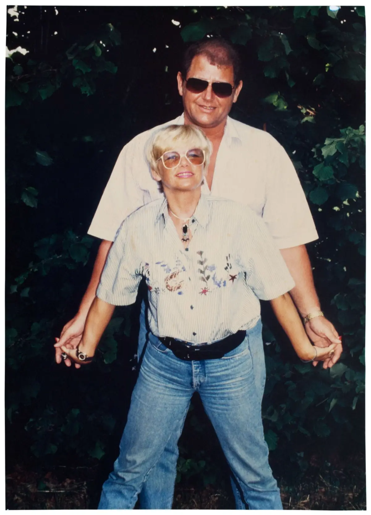 Two adults posing outdoors; foreground adult in striped shirt, behind adult in pale shirt; they hold hands; leafy green background.