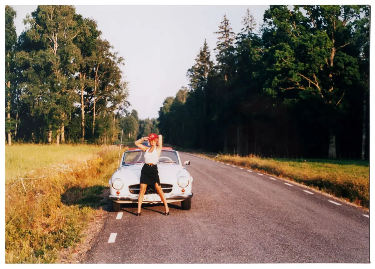 Person in white sleeveless top and black skirt posing beside a classic white convertible on a tree-lined rural road.