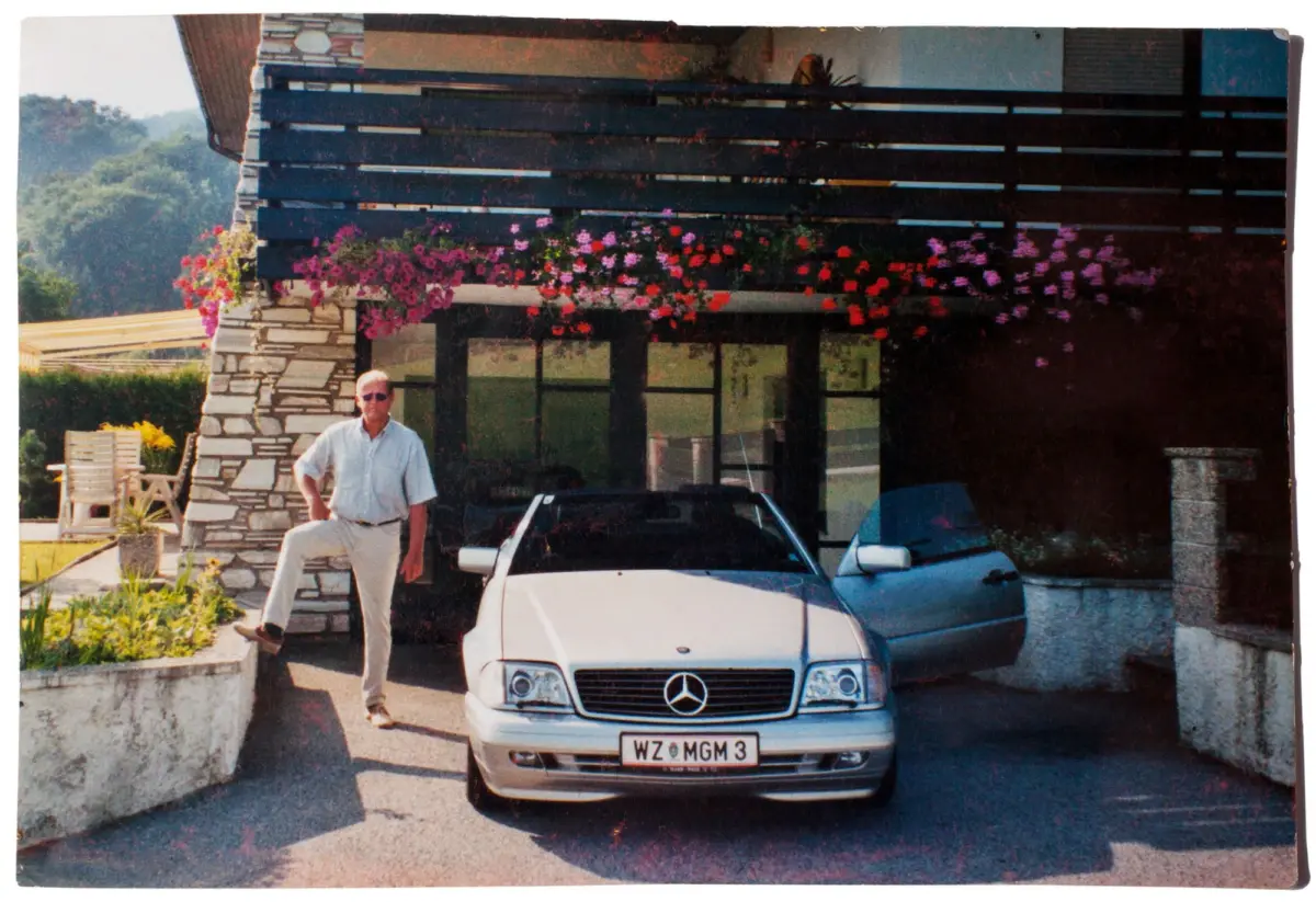 Older man in a light shirt and trousers stands beside a white Mercedes-Benz convertible in a driveway, with a stone house and hanging flowers.