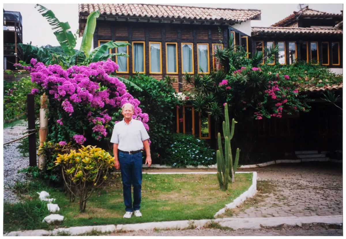 Older man standing in a lush garden in front of a brick house with tiled roof and yellow-framed windows, bougainvillea in bloom.