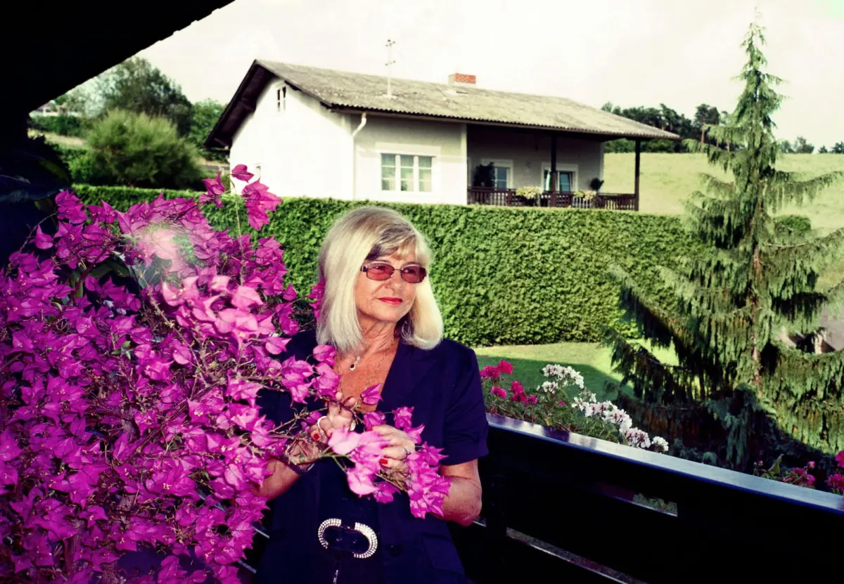 Woman with sunglasses holding pink bougainvillea blossoms in a garden, white house with veranda behind.