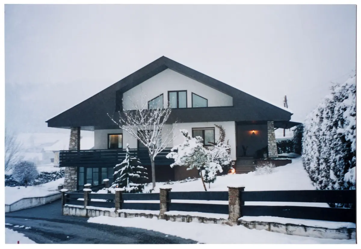 Two-story modern house with a gable roof and large windows, in a snowy yard with bare trees and a wooden fence.