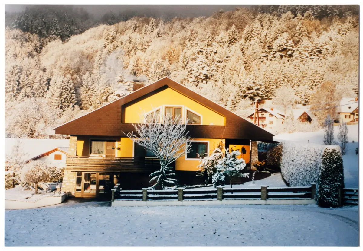 Yellow house with brown roof in a snowy yard; snow-covered evergreen trees on a hillside behind; wooden fence in front.