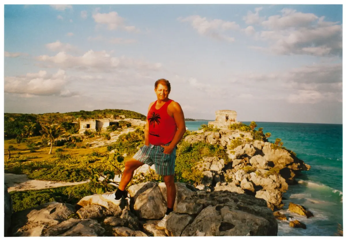 Person standing on rocky coastal outcrop in red palm-tree tank top; turquoise sea and ancient ruins in the background.