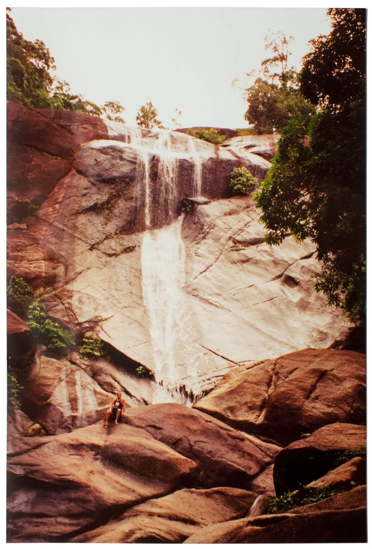 Person sits on a large rock at the base of a cascading waterfall over smooth granite cliffs, surrounded by trees.