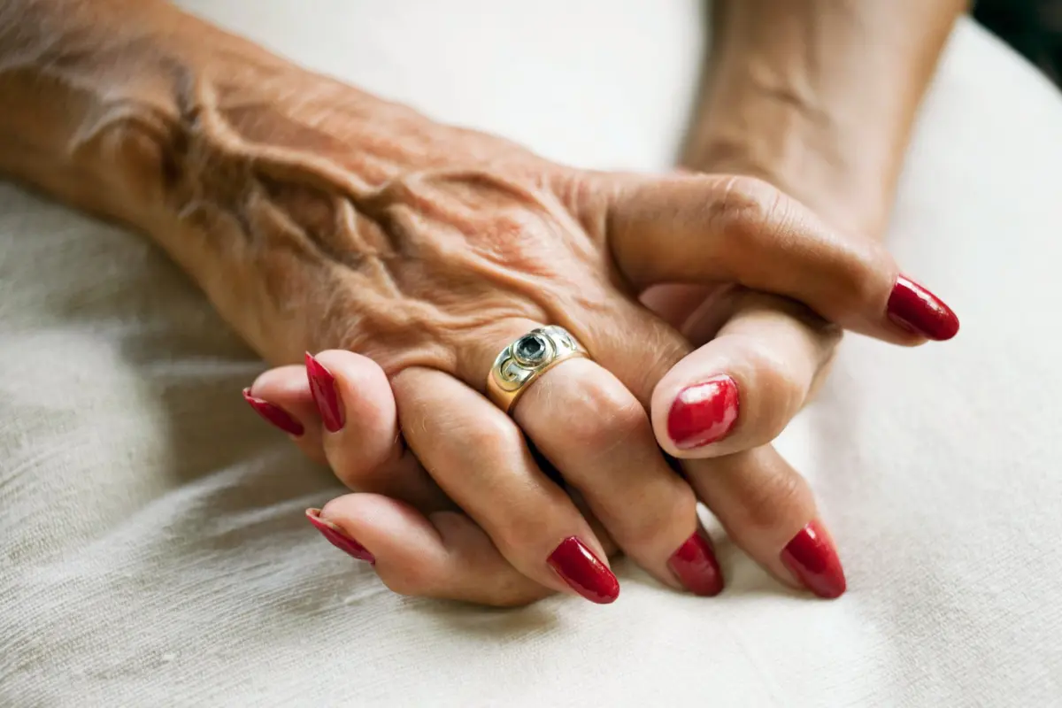 Two elderly hands clasped together; silver ring with a green stone; red-painted nails; resting on light fabric.