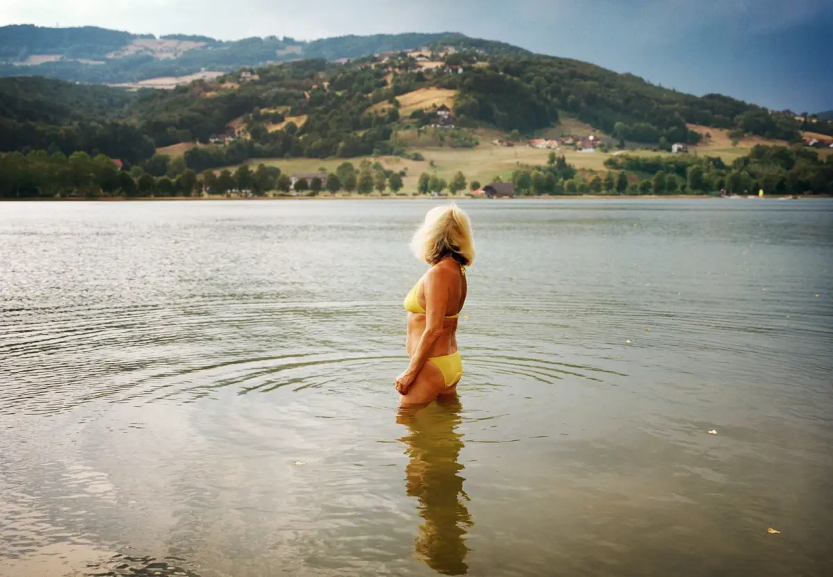 Person in yellow bikini stands waist-deep in lake, with hillside and houses in the distance.