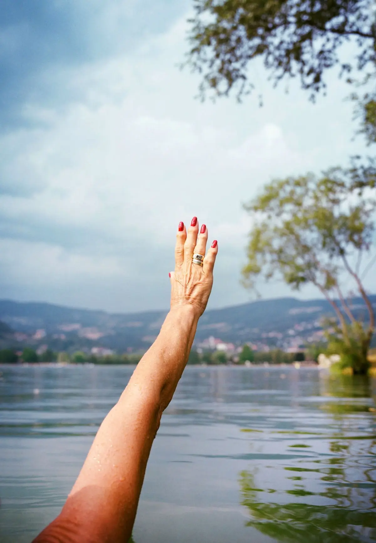 Outstretched arm with red nails and rings raised over a calm lake, with distant mountains and trees in the background.
