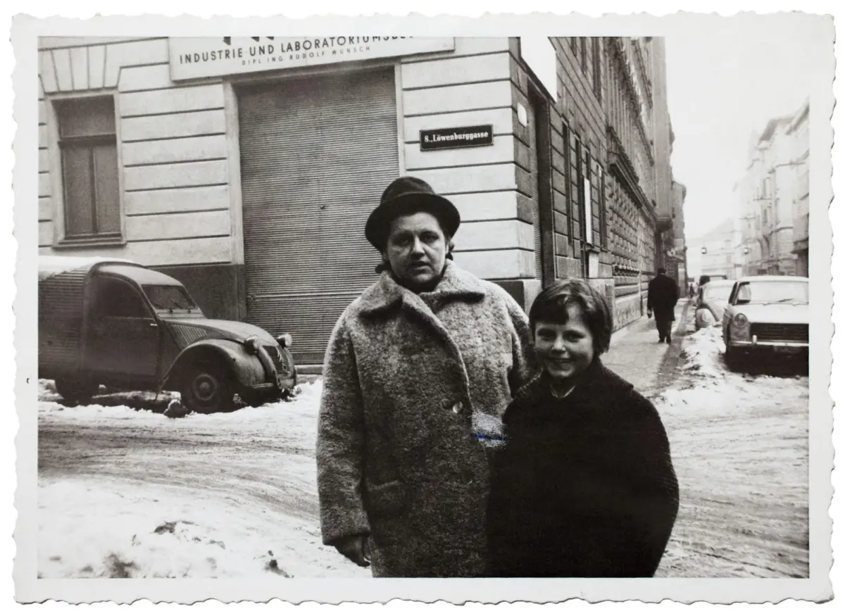 Two pedestrians: an adult in a heavy wool coat and hat, and a smiling child, walk toward the camera on a snowy European city street with vintage cars.