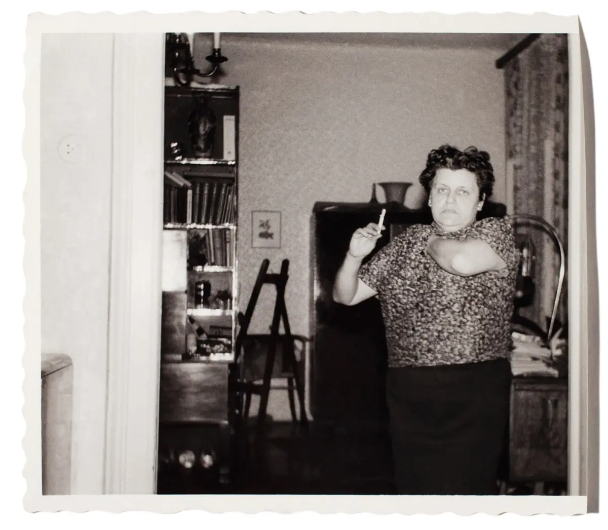 Older person in a patterned dress seated in an armchair, smoking a cigarette, in a mid-century living room.