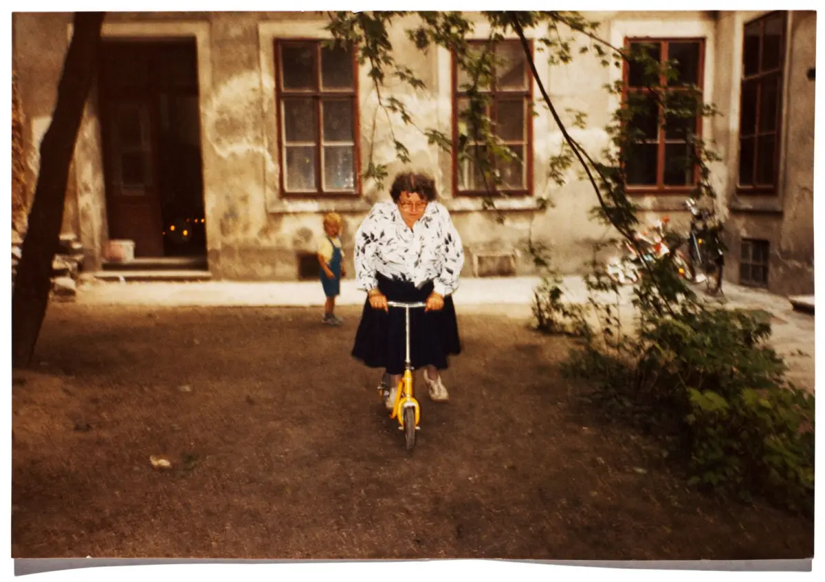 Older person rides a small yellow bicycle toward the camera in a courtyard with peeling walls and climbing vines; a child in blue overalls nearby.
