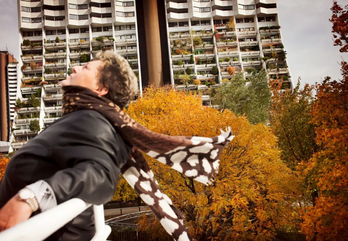 Person in dark jacket and brown scarf leaning on a white railing, gazing upward; urban autumn scene with tall apartment building and orange trees.