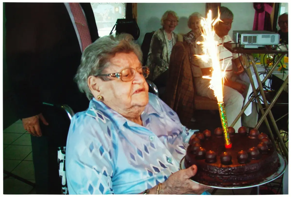 Older woman in a blue patterned blouse seated at a birthday cake with a lit sparkler at a party.