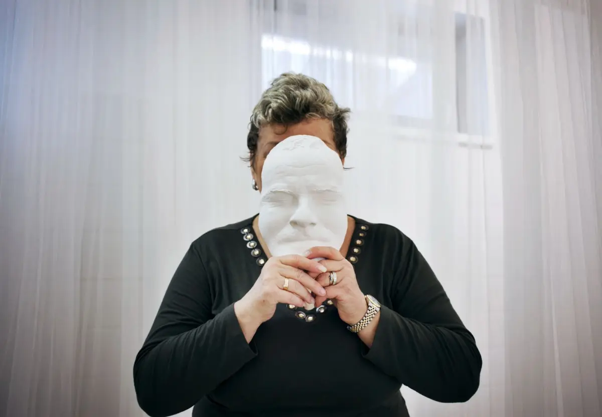 Adult person in a black top with a studded neckline holds a white mask in front of their face indoors behind sheer vertical curtains.