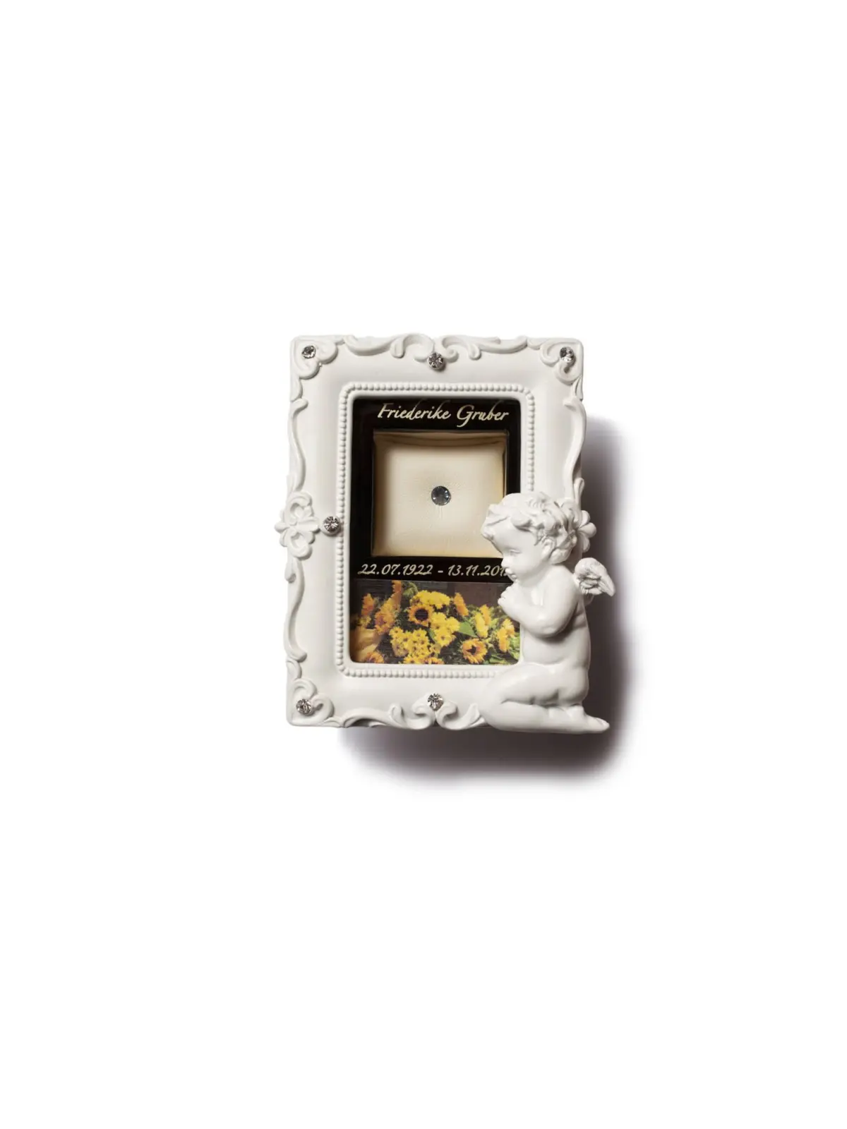 Ornate white frame with a cherub at the lower right, containing a memorial plaque that reads 'Friederike Gruber' with a small jewel on a cream cushion and sunflowers.
