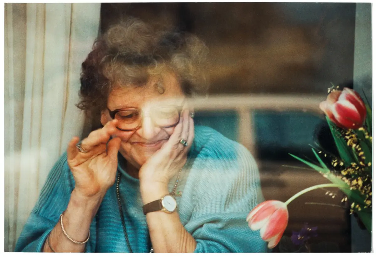 Older person with short curly gray hair and glasses, wearing a blue sweater, smiling with hands on cheeks; tulips and a window in the background.