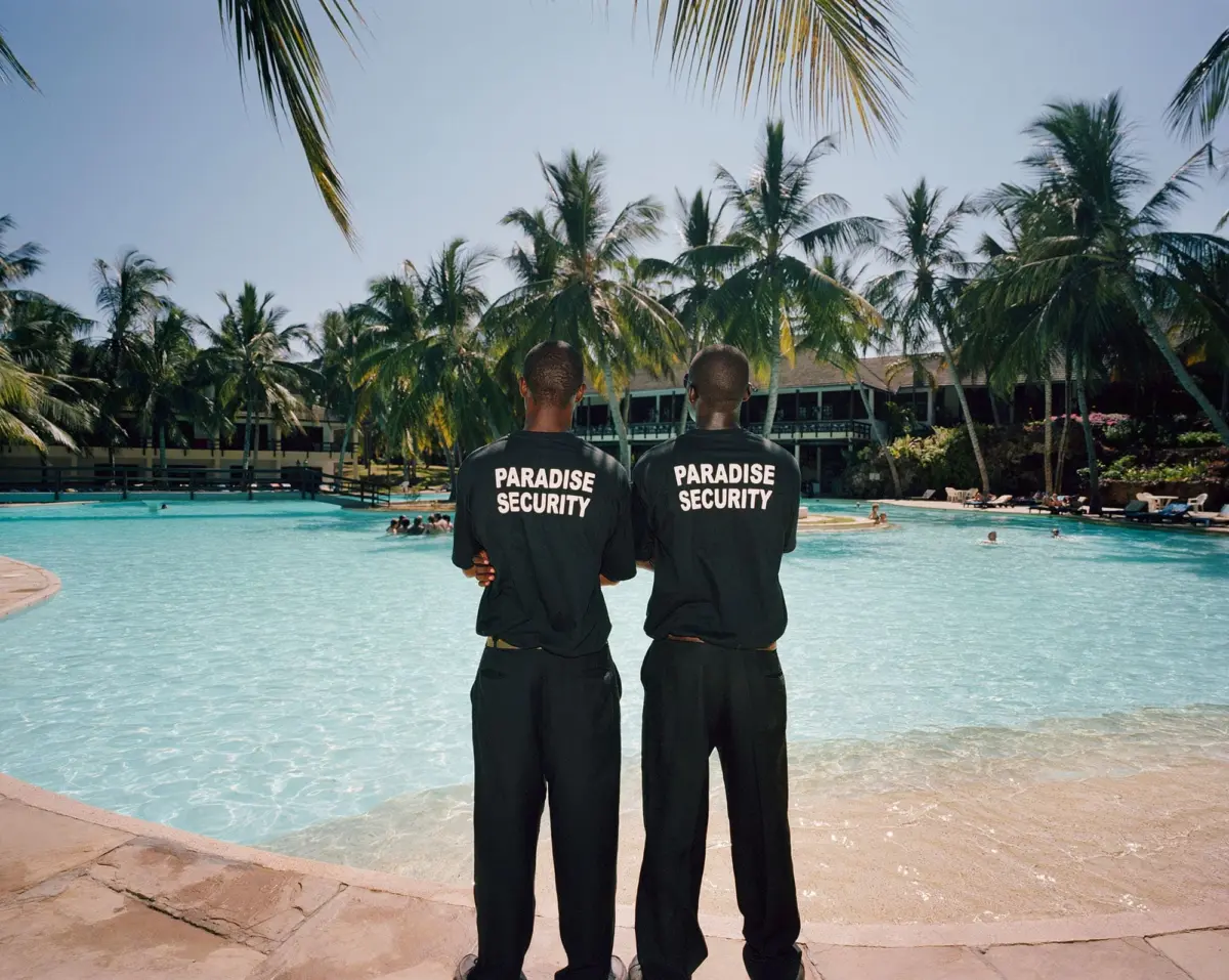 Two security guards wearing Paradise Security shirts stand with arms crossed by a tropical resort pool.