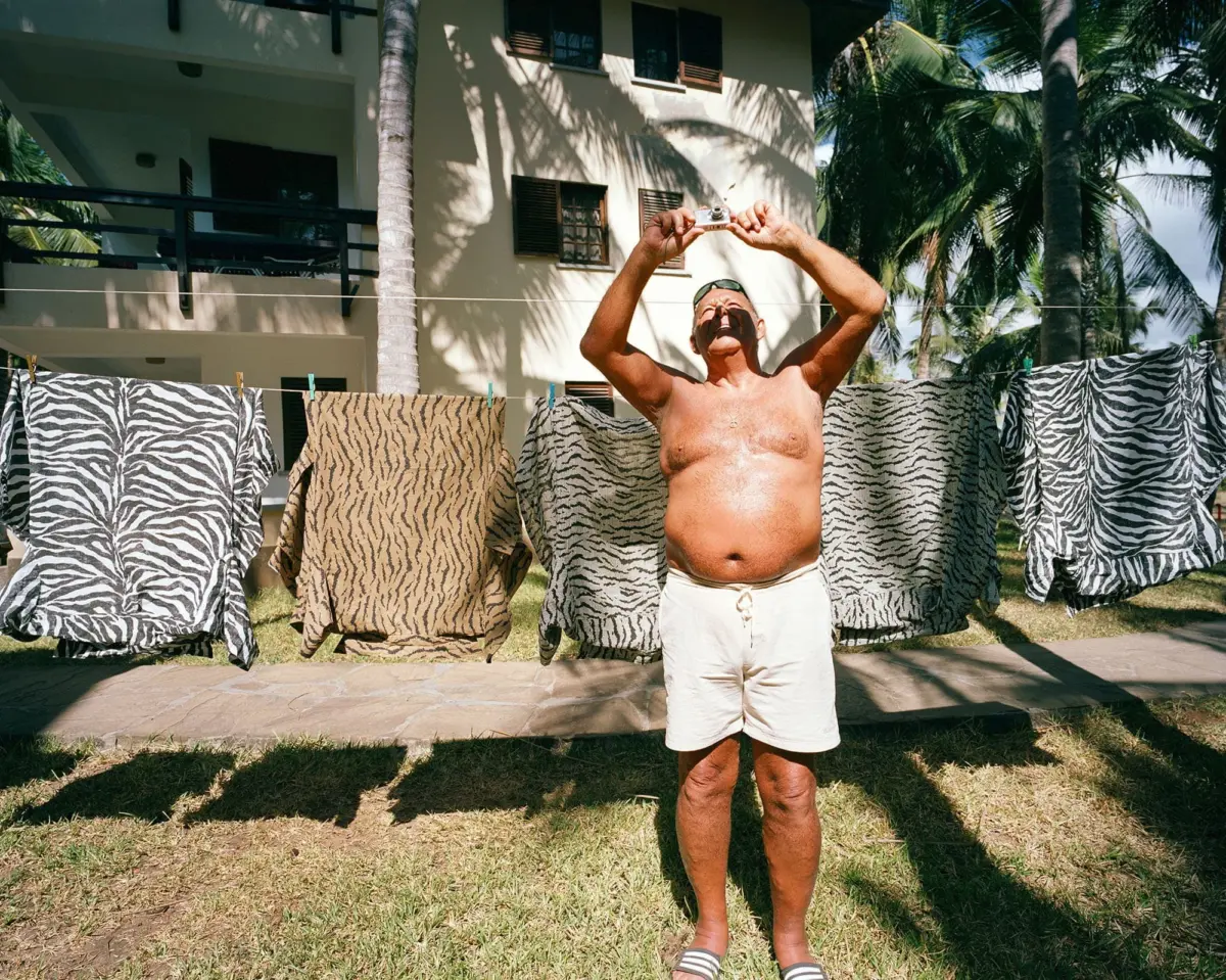 Shirtless adult person raising a small camera above their head in a sunny tropical yard; zebra-pattern towels hanging on a clothesline.
