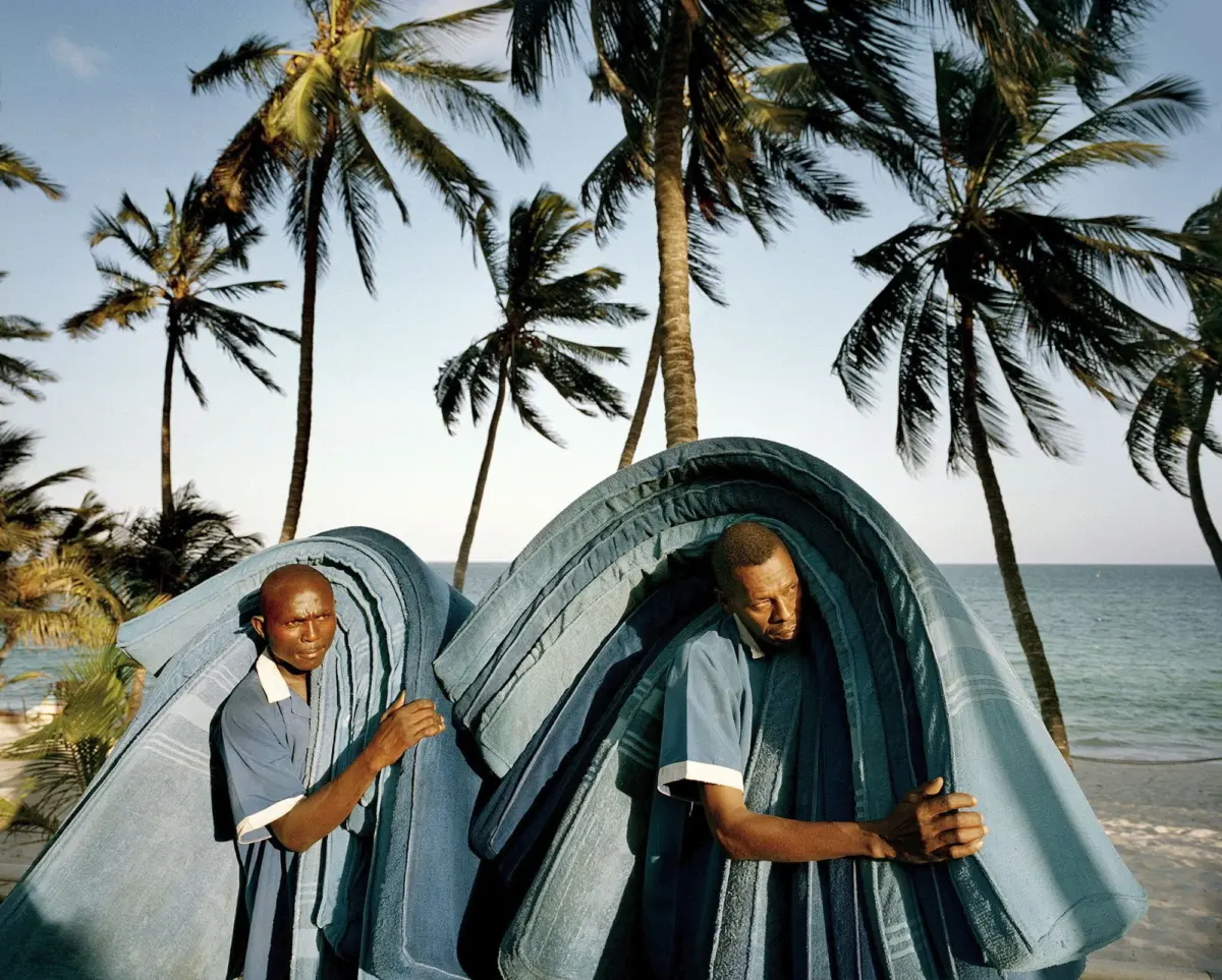 Two people in blue shirts handle oversized blue striped tarps on a palm-lined beach by the sea