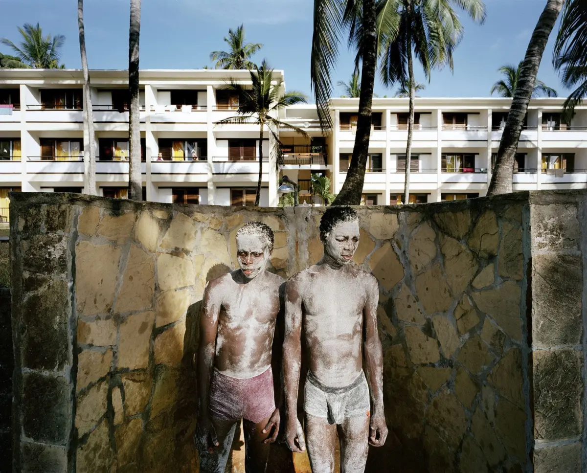 Two shirtless individuals covered in white pigment stand against a stone wall in a tropical resort courtyard with palm trees and a building.