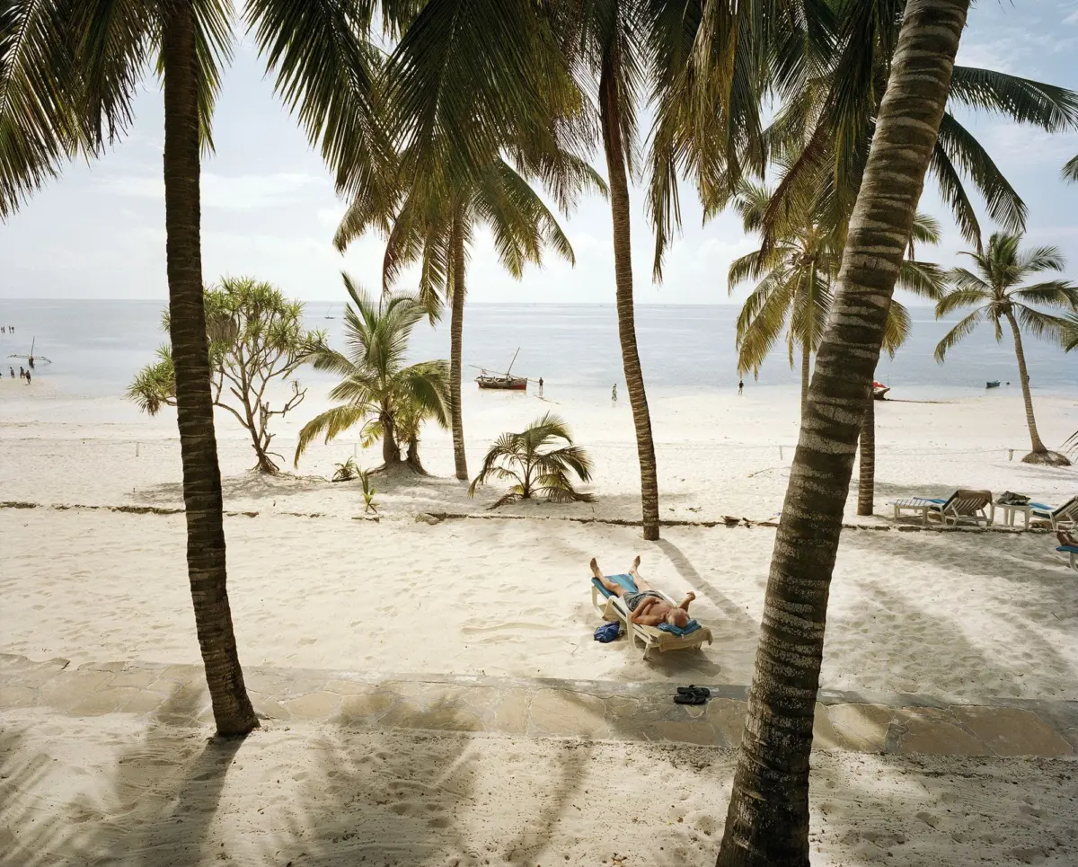 Person sunbathing on a lounge chair on a sandy tropical beach, framed by palm trees with calm sea and distant boat.