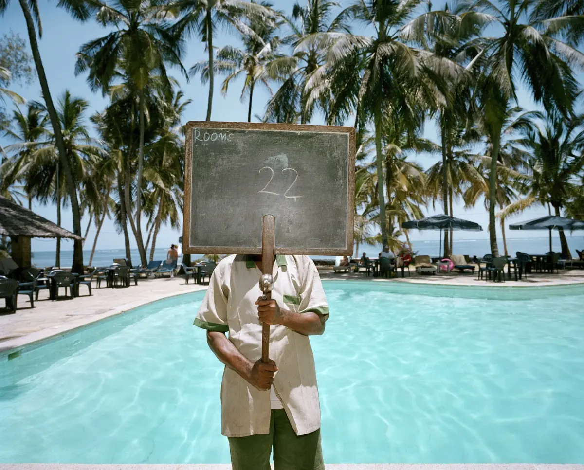 Staff member by a resort pool holds a chalkboard sign showing '22' that blocks their face; palm trees and pool loungers in the background.
