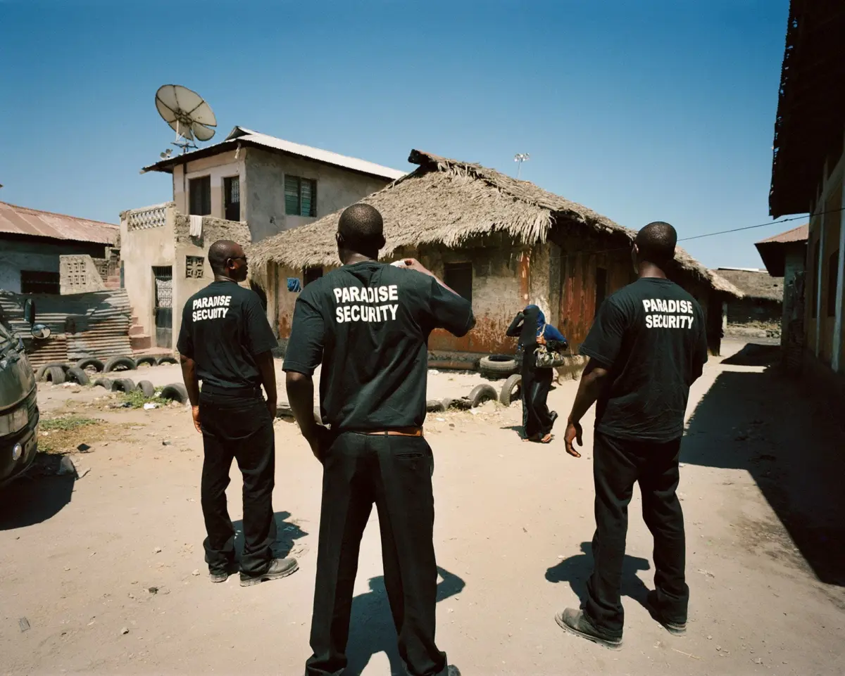 Paradise Security guards stand with backs to camera in a dusty village street; thatched-roof huts and a satellite dish visible.