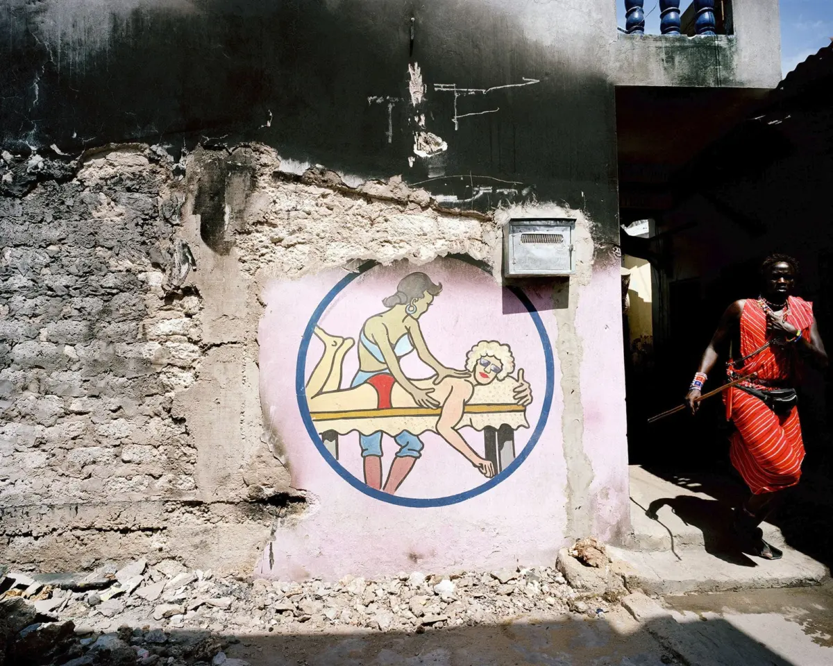 Wall mural inside a pink circle depicts a back massage on a table on a crumbling plaster wall; a person in red striped attire walks by.