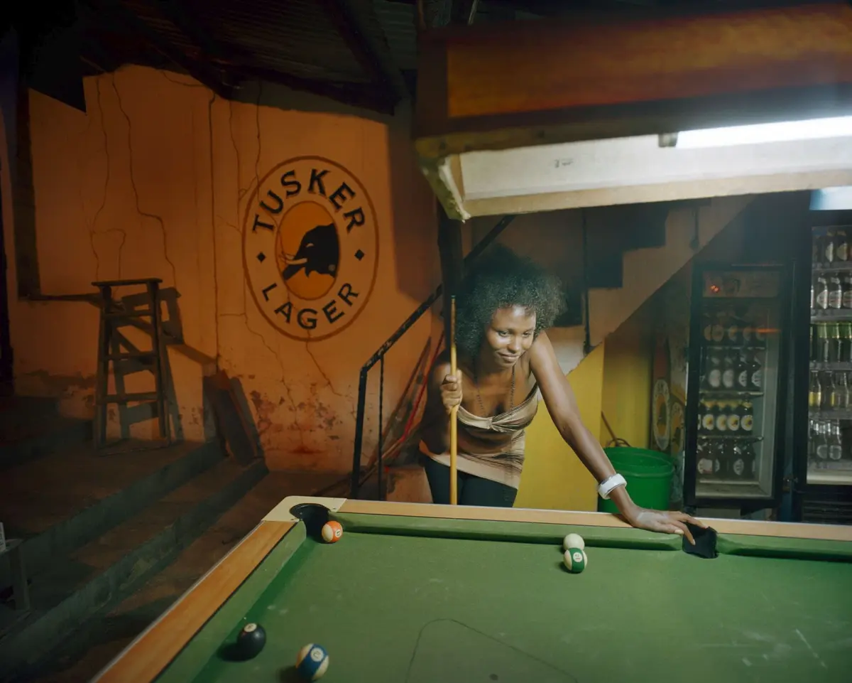 Person leaning over a pool table, cue in hand, in a dim bar with orange wall and Tusker Lager sign; beer fridge on the right.